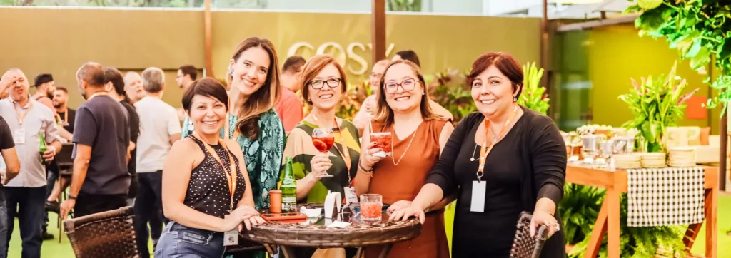 Cinco mulheres atrás de uma mesa pequena e redonda sorrindo para a foto, enquanto acontece um evento com pessoas atrás delas.